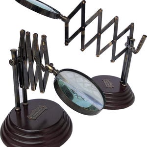 May include: Two magnifying glasses with circular lenses and dark metal frames. Each features an extendable, accordion-style arm mounted on a round, dark brown wooden base. A small rectangular metal plaque is attached to the front of each base.