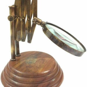 Könnte beinhalten: Eine Lupe im Vintage-Stil mit einem messingfarbenen, verstellbaren Arm und einer runden, klaren Linse. Die Lupe ist auf einem abgestuften Holzsockel mit einem Messingemblem montiert. Das Design erinnert an eine klassische, antike Ästhetik.