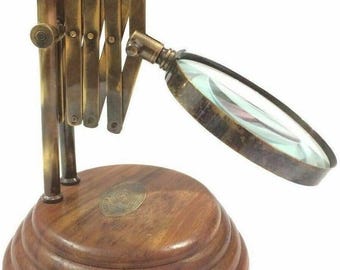 May include: A vintage-style magnifying glass with a brass-colored, adjustable arm and a round, clear lens. The magnifying glass is mounted on a tiered, wooden base with a brass emblem. The design evokes a classic, antique aesthetic.