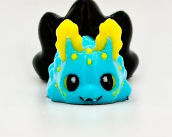 May include: A 3D printed figurine of a sea slug with a bright cyan body, yellow ruffled gills, and yellow spots. The creature has large black eyes and a small mouth. Behind the cyan figure is a larger, solid black, three-lobed shape.