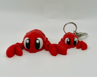 May include: Two bright red, 3D-printed crab figures with large, cartoon-style eyes featuring white pupils and black irises. The larger crab is positioned on the left, while the smaller crab on the right is attached to a silver metal keyring.
