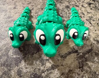 May include: Three articulated green alligator figures with segmented bodies and textured backs. Each figure features large white eyes with black pupils. The central alligator is the largest, flanked by two smaller alligator figures on each side.