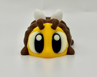 May include: A 3D-printed bee figurine featuring a bright yellow face, large black and white eyes, and brown body segments. The insect has two small white antennae on top and a rounded, layered structure with visible horizontal print lines.