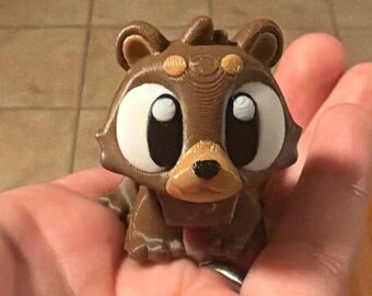 May include: A small, brown, 3D-printed figurine of a stylized bear with large, white, oval-shaped eyes, black pupils, and a light brown snout with a black nose. The bear has rounded ears and visible horizontal layer lines from the printing process.