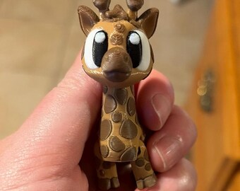 May include: A small 3D printed giraffe figurine featuring a tan body with brown spots, large white and black eyes, and two small ossicones on top of its head. The surface of the plastic figure shows visible horizontal layer lines from the printing process.