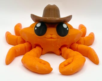 May include: A bright orange crab figurine featuring large black eyes with white highlights, wearing a brown cowboy hat. The crab has multiple articulated legs extending from its body and two front claws positioned forward on a plain white background.