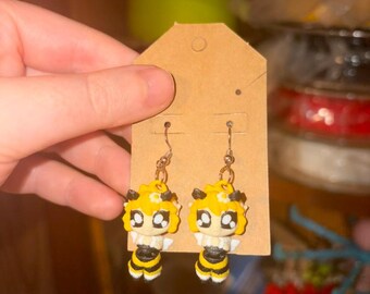 May include: A pair of dangle earrings featuring sculpted bee characters with yellow hair, large black and white eyes, and black and yellow striped bodies. The earrings are attached to a brown rectangular cardboard display card with a hole at the top.