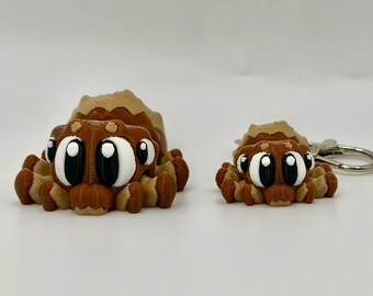 May include: Two 3D printed spider figurines in brown and tan with large white and black eyes. The larger spider is a standalone figure, while the smaller spider is attached to a silver metal keychain ring, both featuring a layered texture.