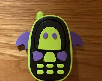 May include: A lime green and black toy mobile phone with purple bat-like wings on the sides. The device features a small antenna, a screen area with two purple eyes, and a keypad with lime green buttons set against a black background.