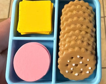May include: A light blue plastic divided tray containing a stack of yellow square slices, a pink circular disc, and a vertical row of brown scalloped-edge round crackers with small white dots on the surface of the top cracker.