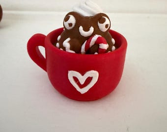 May include: A miniature red mug featuring a white heart outline on the front. Inside the mug sits a brown frog-like figure with white eyes and a mouth, topped with white whipped cream and a small red and white striped candy cane accessory.