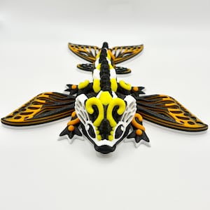 以下が含まれることがあります： 3Dプリントされたドラゴンの置物。翼と胴体は層状になっており、黒、白、黄色、オレンジの配色です。翼は黒とオレンジの層になっており、胴体はセグメント化されたデザインです。