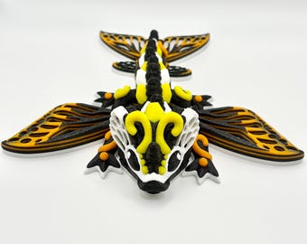 May include: A 3D printed articulated dragon figurine featuring a black, white, and yellow color scheme. The creature has intricate wing patterns, a segmented body, and detailed head ornamentation with curved yellow accents and black eyes.