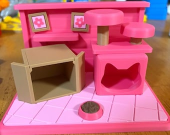 May include: A miniature pink cat play set featuring a cat tree with two platforms, a cat-shaped entrance to a cubby, a tan cardboard-style box, and a small pink bowl with brown contents. The back wall displays two small framed flower pictures.