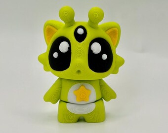 May include: A lime green 3D printed figurine featuring a creature with two antennae, three eyes, and large black eye patches. The character has yellow inner ears, a yellow star on its chest, and a white belly patch, all with visible layer lines.