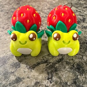 Könnte beinhalten: Zwei 3D-gedruckte Erdbeerfrosch-Figuren. Jeder Frosch ist gelb mit weißem Bauch, braunen Augen und einer grünen Blattkrone, die eine rote Erdbeere mit gelben Samen trägt. Einer hat einen Schlüsselanhänger.