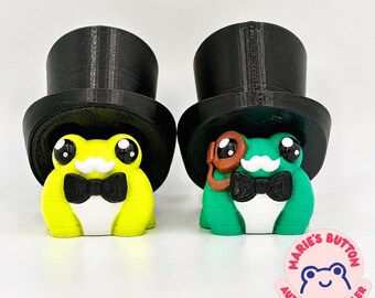 May include: Two 3D-printed frog figurines wearing black top hats, white mustaches, and black bow ties. The left frog is lime green, and the right frog is emerald green with a brown monocle. A circular logo reads Marie's Button Authorized Seller.