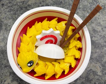 May include: A white bowl with a red interior containing a yellow articulated creature with large black and white eyes. The bowl includes a white and red fish cake slice, a smooth white dome, and two brown textured sticks resembling chopsticks.