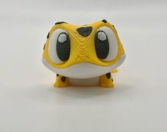 May include: A small, 3D-printed figurine of a leopard gecko with a bright yellow body, a white underbelly, and black spots. The creature features large, expressive eyes with black pupils and white highlights, and a textured surface finish.