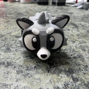 Peut inclure: Figurine de raton laveur imprimée en 3D, dans des tons de gris, de noir et de blanc. Le raton laveur a de grands yeux expressifs, un masque noir et un museau blanc. Les oreilles sont à bout noir et le corps a un design épineux. C'est un objet décoratif fantaisiste.