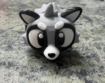 May include: A 3D printed raccoon figurine featuring a gray, black, and white color scheme. The character has large, prominent eyes with black outlines, a small black nose, and pointed ears. The surface shows visible horizontal layer lines.
