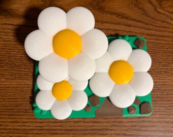 May include: A decorative object featuring three white flowers with bright yellow centers, mounted on a rectangular green base. The base includes small brown rock-like accents and green leaf details, all rendered with a visible 3D printed texture.