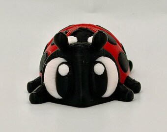 May include: A 3D printed ladybug figurine featuring a black head and legs, a red back with black spots, and large white eyes with black pupils. The surface shows visible horizontal layer lines from the printing process, creating a textured finish.