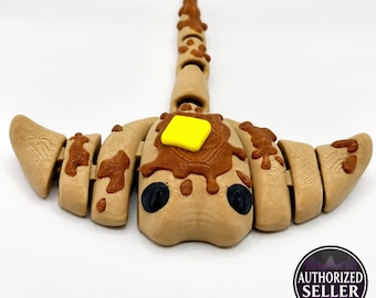 May include: A tan, articulated, segmented creature figure resembling a pancake with a bright yellow square butter pat on top and brown syrup drips. The figure has two dark eyes and a long, segmented tail extending from the center back.