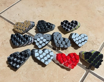 May include: A collection of eleven heart-shaped objects, each constructed from a cluster of small hexagonal segments. The hearts feature various color combinations, including solid red, black, gray, white, and tan, as well as striped patterns.