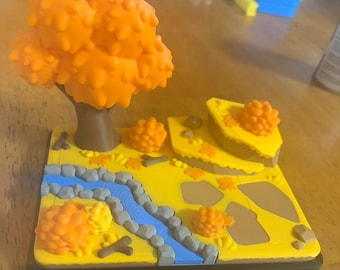 May include: A 3D printed landscape diorama featuring a brown tree with orange foliage, a blue winding river bordered by gray stones, yellow ground terrain, brown rock formations, and small orange bushes on a square black base.