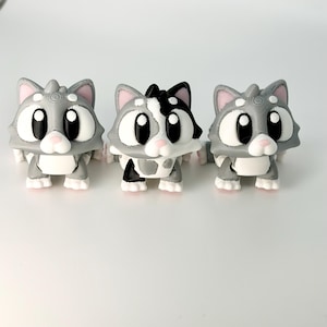 May include: Three 3D-printed cat figurines. Two are gray with white and pink accents, and one is black and white. The cats have large, round eyes and are standing upright. The figurines are on a white surface.