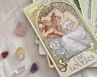 May include: A tarot card titled "The Lovers" with an illustration of two figures embracing, framed by gold detailing and white roses. Several colourful gemstones, including rose quartz, are scattered around the cards.