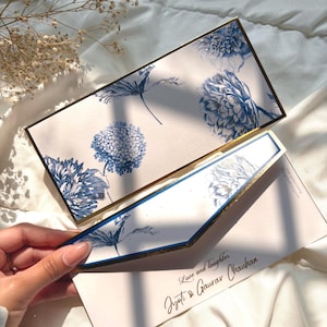 Puede incluir: Elegante juego de invitaciones de boda con diseños florales azules sobre fondo blanco, con ribete dorado. La invitación incluye el texto "Love and laughter" y los nombres "Jyoti & Gaurav Chauhan".