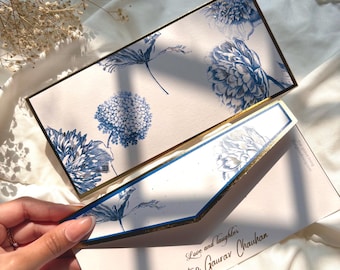 May include: Elegant wedding invitation set featuring blue floral designs on a white background, with gold trim. The invitation includes the text "Love and laughter" and the names "Jyoti & Gaurav Chauhan".