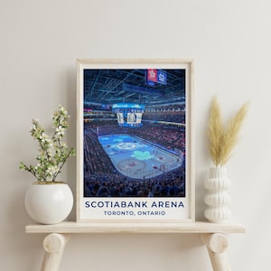 Può includere: Una stampa incorniciata che mostra una vista interna della Scotiabank Arena a Toronto, Ontario, con una pista da hockey caratterizzata da un grande logo blu a forma di foglia d'acero al centro, circondata da spalti e un tabellone luminoso.