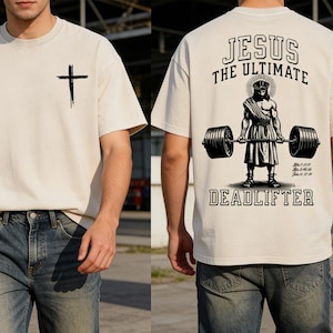 以下が含まれることがあります： 前面に黒い十字架が付いたクリーム色のTシャツ。背中には「JESUS THE ULTIMATE DEADLIFTER」の文字と、ウェイトリフティングをするイエスのグラフィックが描かれています。柔らかい素材でクルーネックです。