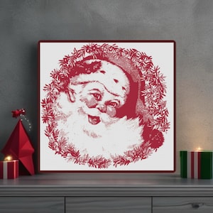 May include: Red and white Christmas art print featuring Santa Claus with a wreath. The vintage-style illustration shows Santa's face surrounded by a wreath of leaves. The print is framed in a dark red border and is displayed on a gray surface.