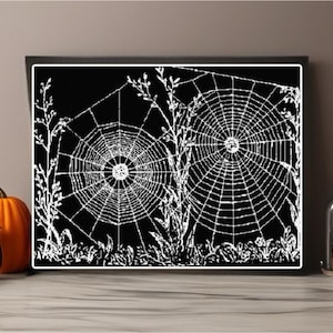 May include: A framed black and white print featuring two detailed spiderwebs. The artwork is surrounded by white foliage and grass, evoking a Halloween theme. Decorative pumpkins and a jar with cookies are in the foreground.