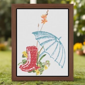 May include: A framed cross-stitch design showing a red polka-dot wellington boot, a light blue umbrella, and yellow flowers. The artwork is set against a white background and framed in a brown wooden frame. The design evokes a spring or garden theme.