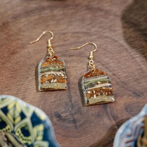 May include: A pair of gold-coloured dangle earrings. Each earring has a clear, rectangular resin pendant with layers of dried plant material and gold leaf. The earrings hang from gold-coloured ear wires.
