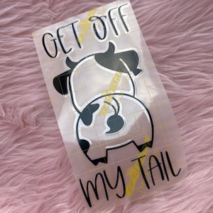 May include: A clear sticker with a cartoon cow design, featuring black and white spots. The text "GET OFF MY TAIL" is written in black, with yellow lightning bolt accents. The sticker is on a pink, fluffy surface.