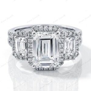 May include: A three-stone engagement ring featuring an emerald-cut central diamond, flanked by two smaller emerald-cut stones, all encircled by a halo of smaller diamonds. The ring is set in a white metal band.