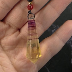May include: A faceted, pointed pendant made of translucent yellow and purple banded stone. The pendant is suspended from a black cord featuring a single small, round, red bead and a tiny gold-toned accent bead above the stone's top point.