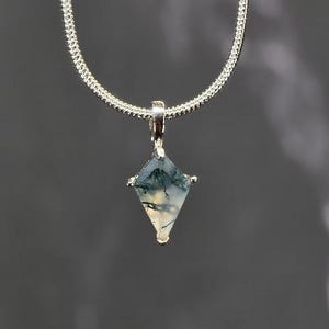 Puede incluir: Un collar de plata con un colgante en forma de diamante. El colgante presenta una piedra natural con patrones verdes y blancos. La cadena es de plata fina y texturizada. El colgante est&aacute; suspendido de una peque&ntilde;a fianza de plata.