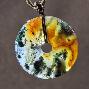 May include: A circular pendant with a central hole, showcasing a blend of colors including yellow, orange, green, and white. The pendant is suspended by a black cord and a green braided necklace.
