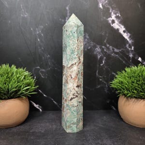 May include: A tall, six-sided crystal tower featuring a polished surface with a marbled pattern of sage green, white, and dark brown mineral inclusions. The stone has a pointed, faceted top and a flat base, displaying natural color variations.