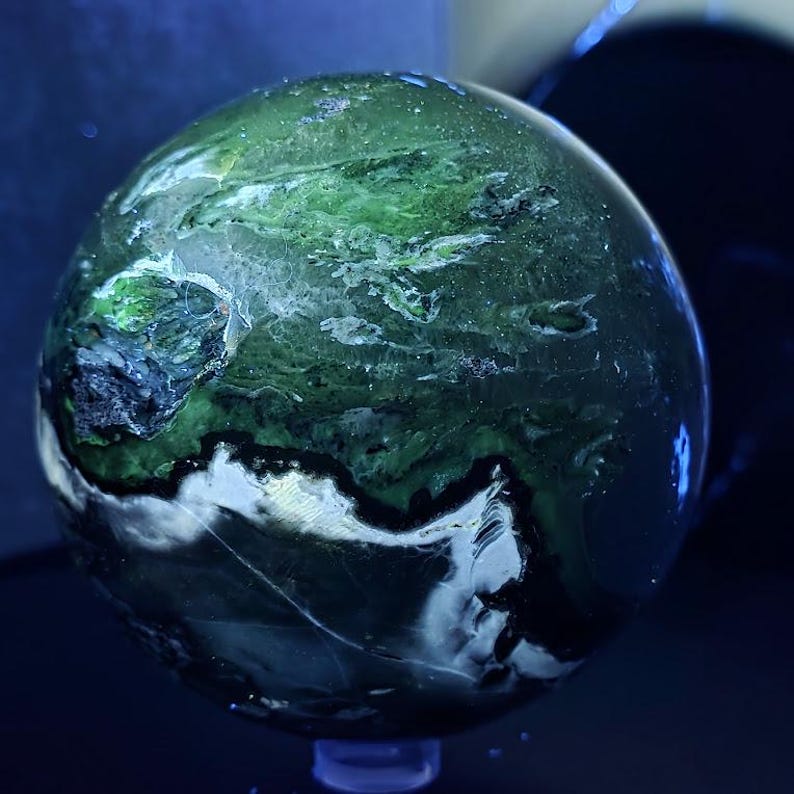 Peut inclure: Une pierre pr&eacute;cieuse sph&eacute;rique polie avec un motif tourbillonnant de vert, de blanc et de noir. L'orbite est plac&eacute;e sur un fond sombre, mettant en valeur ses couleurs et textures naturelles. C'est un objet d&eacute;coratif.