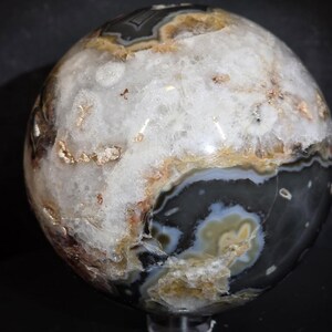 May include: A polished, spherical stone with a mix of colors. The stone features a white, crystalline center surrounded by bands of gray, brown, and tan hues. The surface has a glossy finish, highlighting the natural patterns.