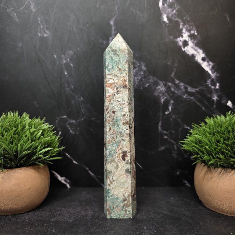 May include: A tall, six-sided crystal tower featuring a mottled pattern of sage green, cream, and dark brown mineral inclusions. The stone has a smooth, polished surface that tapers to a pointed apex, showcasing natural variations in color and texture.