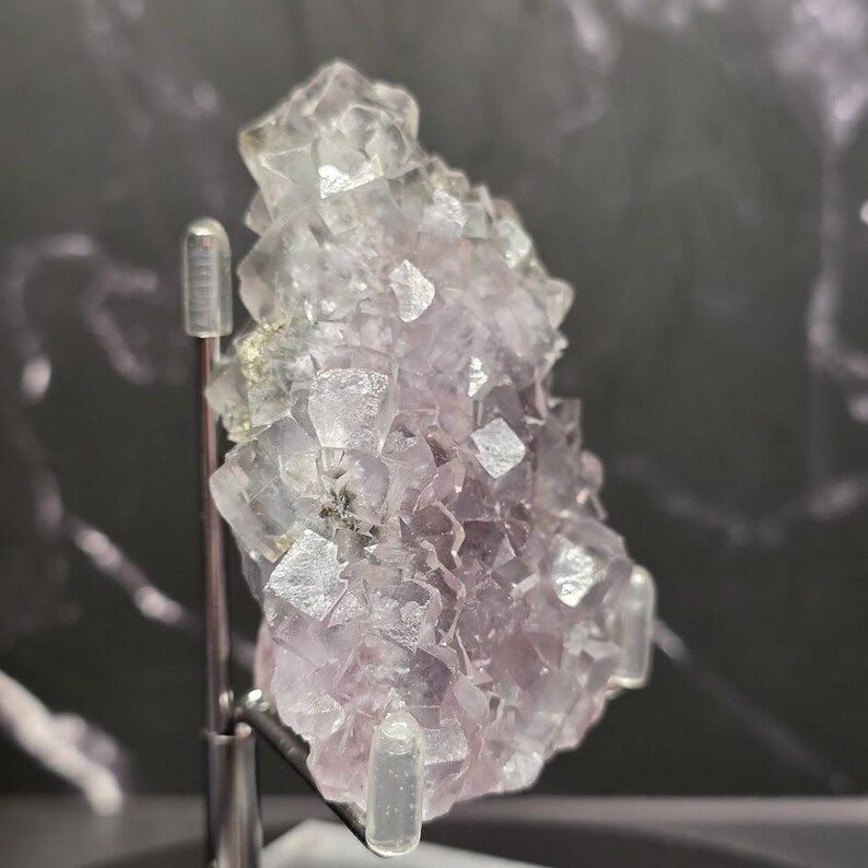 Puede incluir: Un grupo de cristales de fluorita, que muestra una mezcla de estructuras cristalinas de color p&uacute;rpura p&aacute;lido y transparente. La muestra mineral est&aacute; montada sobre un soporte plateado, con un fondo oscuro.